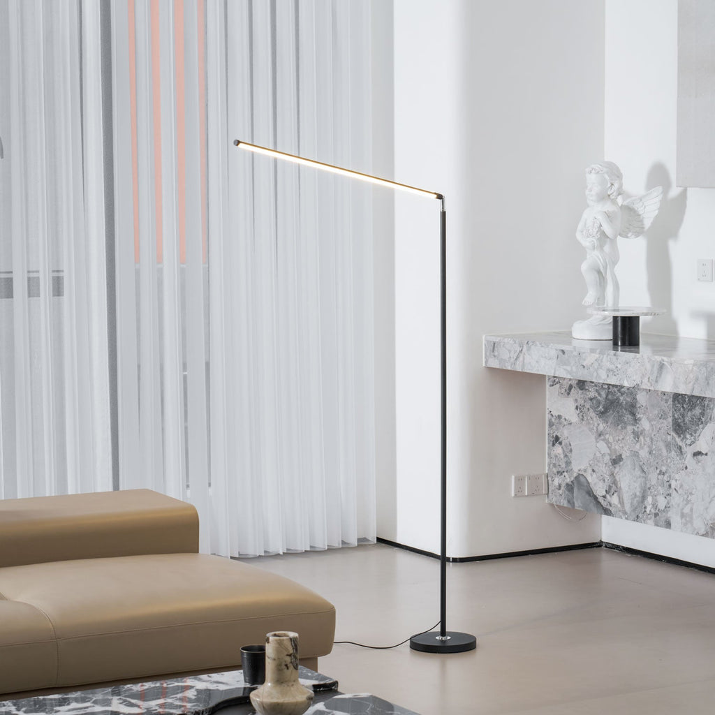 Black Clérachic floor lamp with a lit bar, in a bright room with a stone console, a small statue and a tan sofa with a cord on the floor.