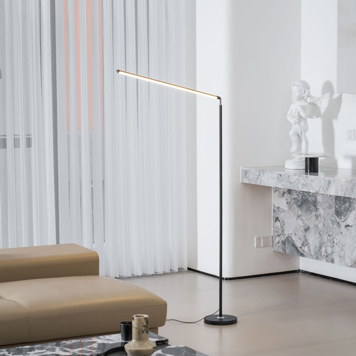 Black Clérachic floor lamp with a lit bar, in a bright room with a stone console, a small statue and a tan sofa with a cord on the floor.
