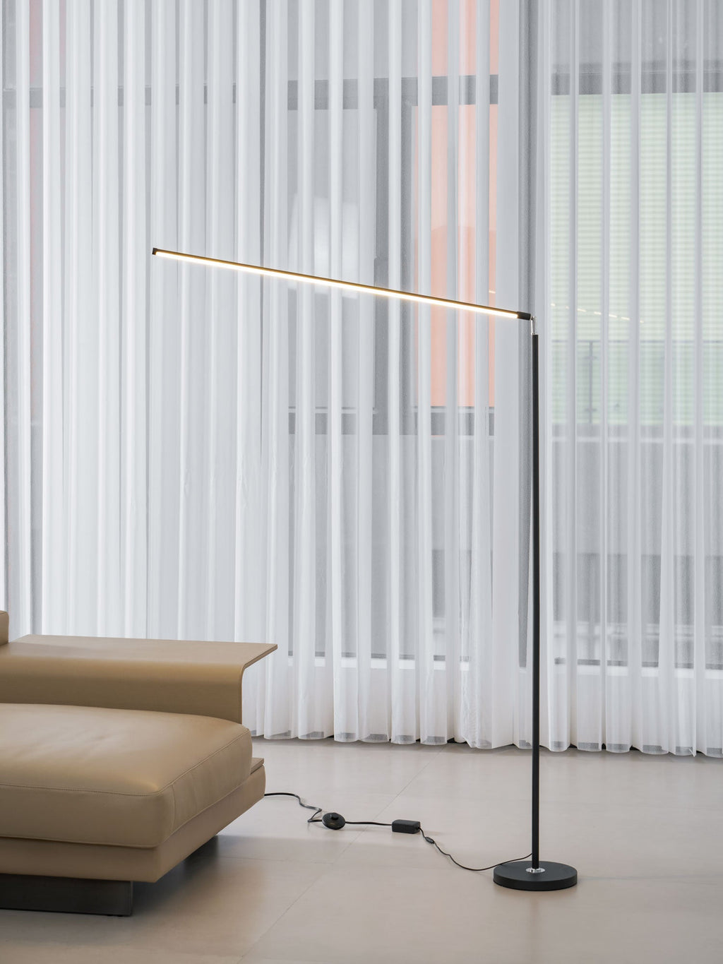 Black Clérachic floor lamp seen from the sofa side, its light bar tilted slightly, with the inline switch lying on the floor.