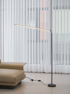 Black Clérachic floor lamp seen from the sofa side, its light bar tilted slightly, with the inline switch lying on the floor.