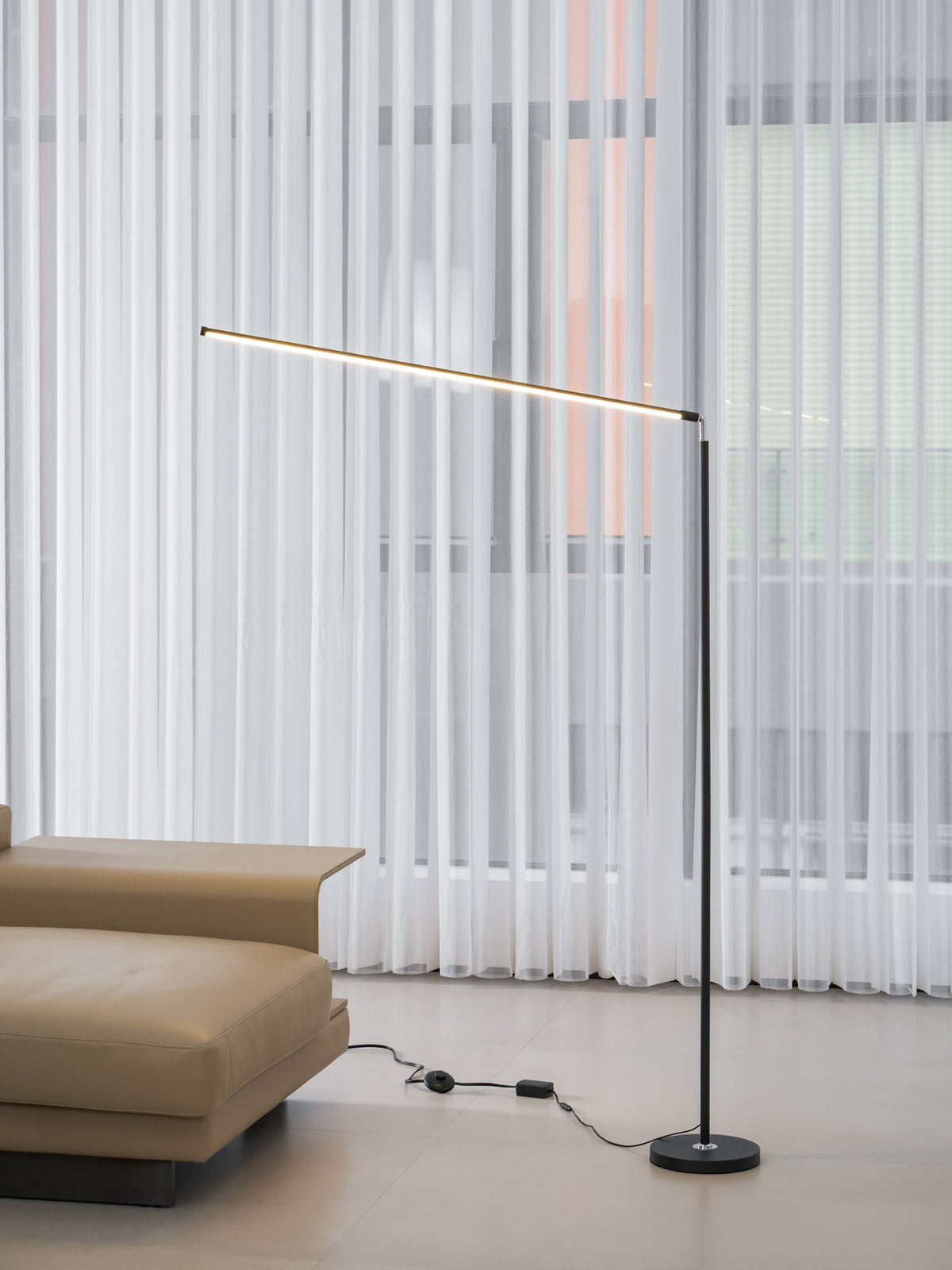 Black Clérachic floor lamp seen from the sofa side, its light bar tilted slightly, with the inline switch lying on the floor.