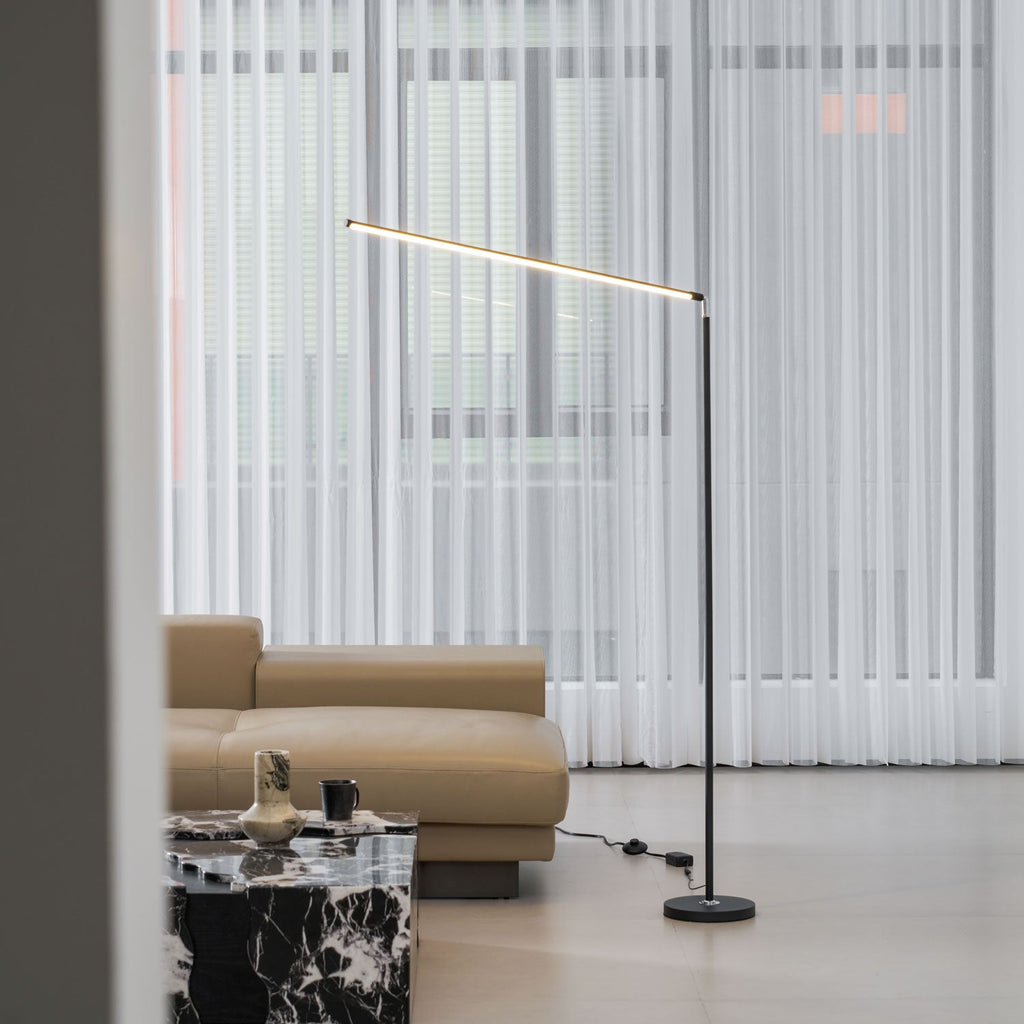 This Clérachic floor lamp, with its pole and round base in black, stands by sheer curtains, a tan sofa and a marble-top table.