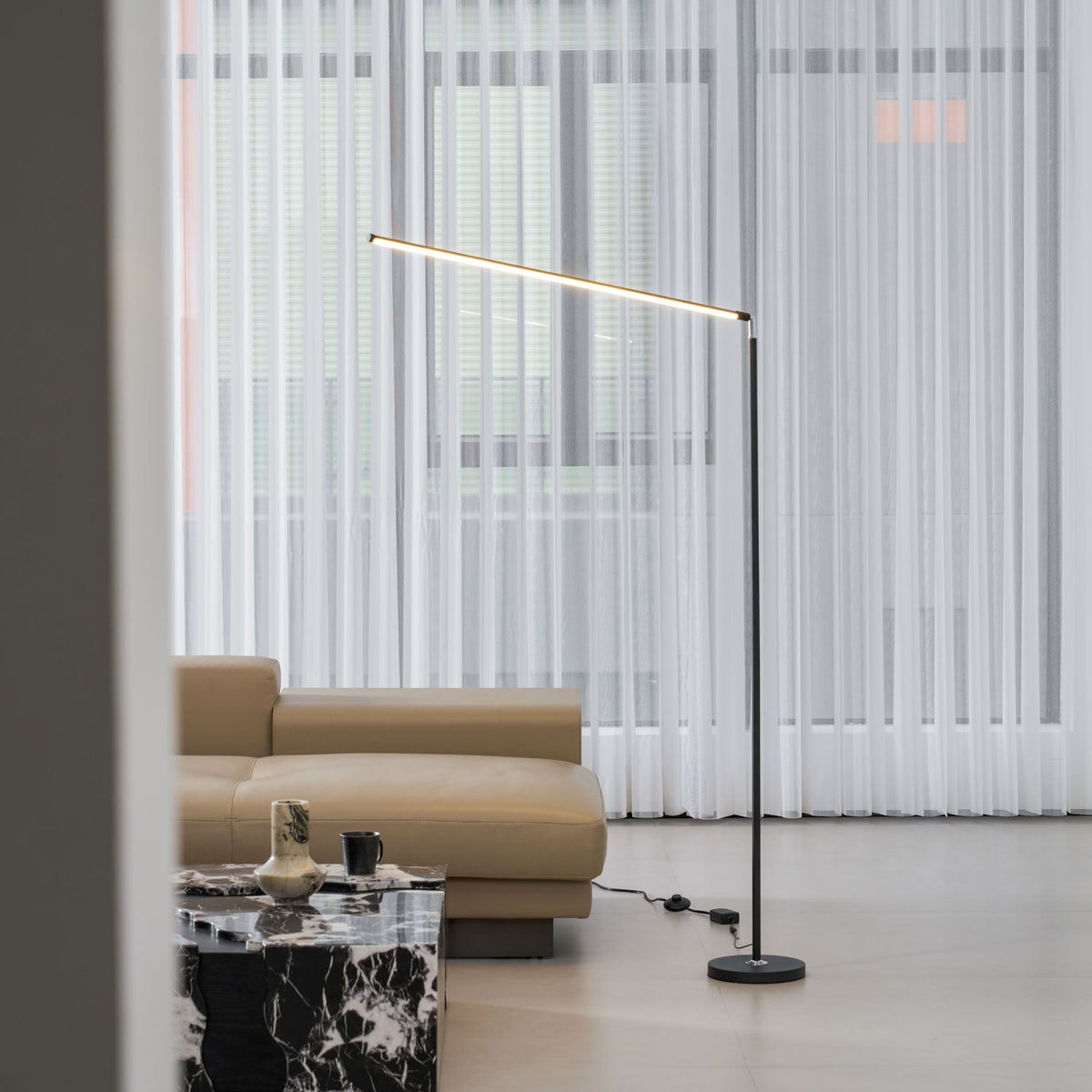 This Clérachic floor lamp, with its pole and round base in black, stands by sheer curtains, a tan sofa and a marble-top table.