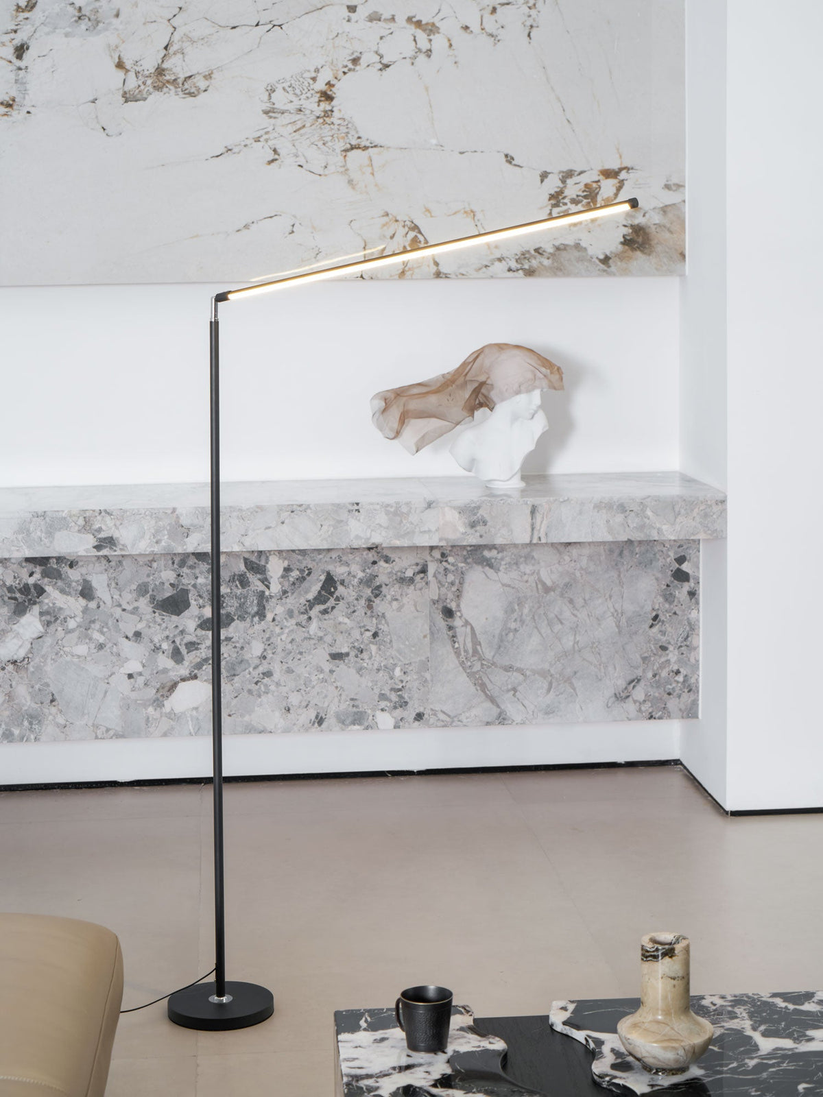 This Clérachic floor lamp lights a marble-look wall behind a stone console with a small sculpture, its bar tilted upwards.