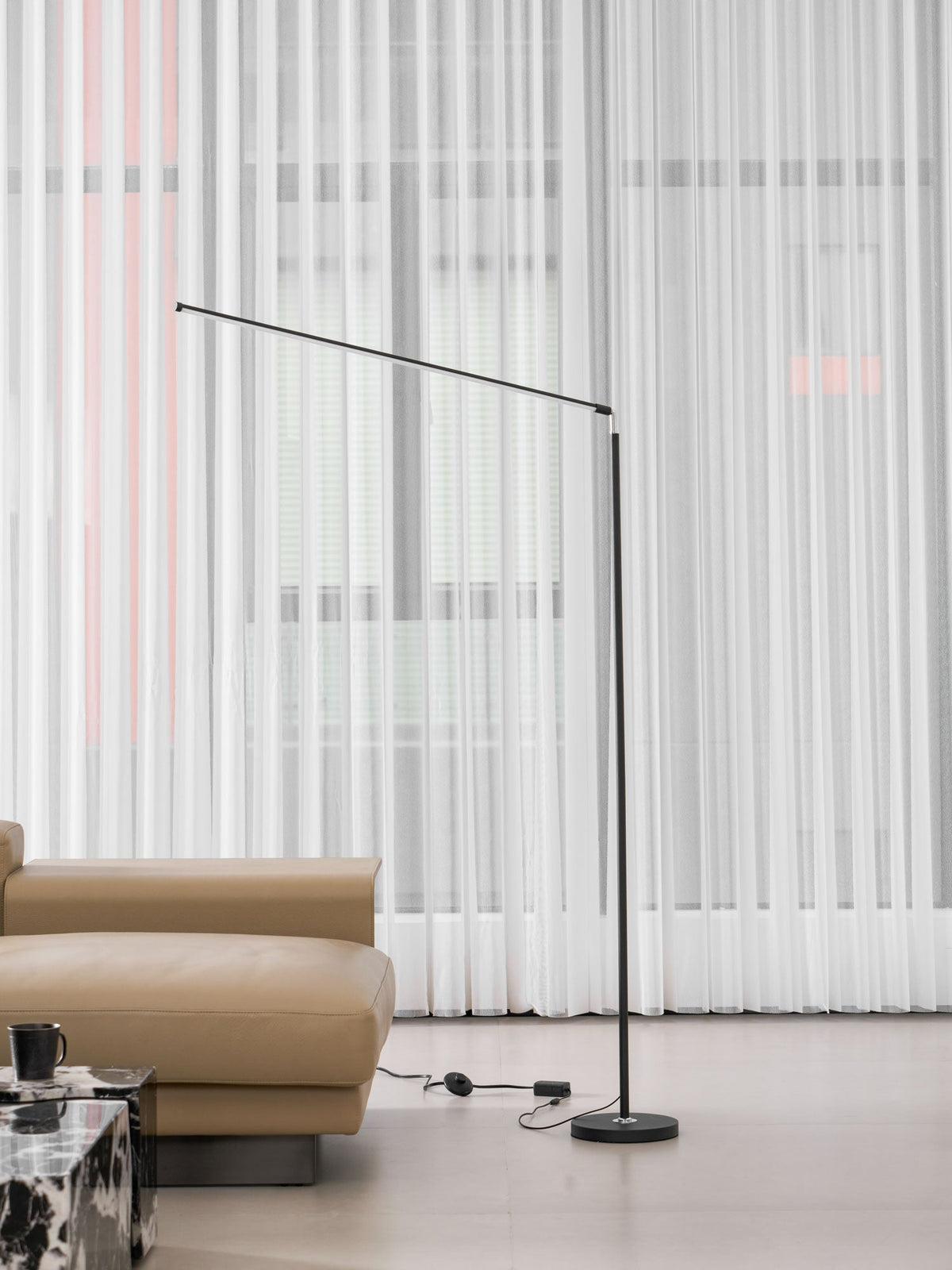 Black Clérachic floor lamp with the bar held out to the left, in front of sheer curtains and a tan sofa, with its cord on the floor.