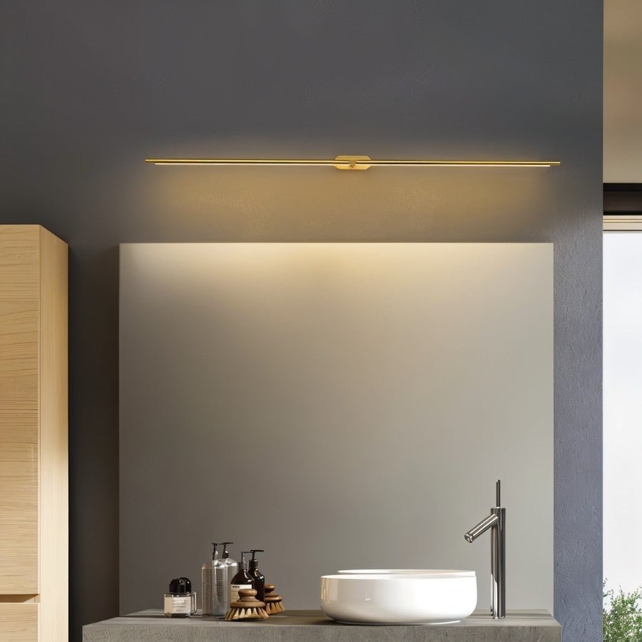 Clérachic slim gold bar light centred above a mirror on a slate-grey wall, with a white basin on a stone shelf below.