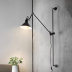 A Clérachic black wall light with a lit cone shade, mounted on a textured concrete-look wall with a white flower vase below.