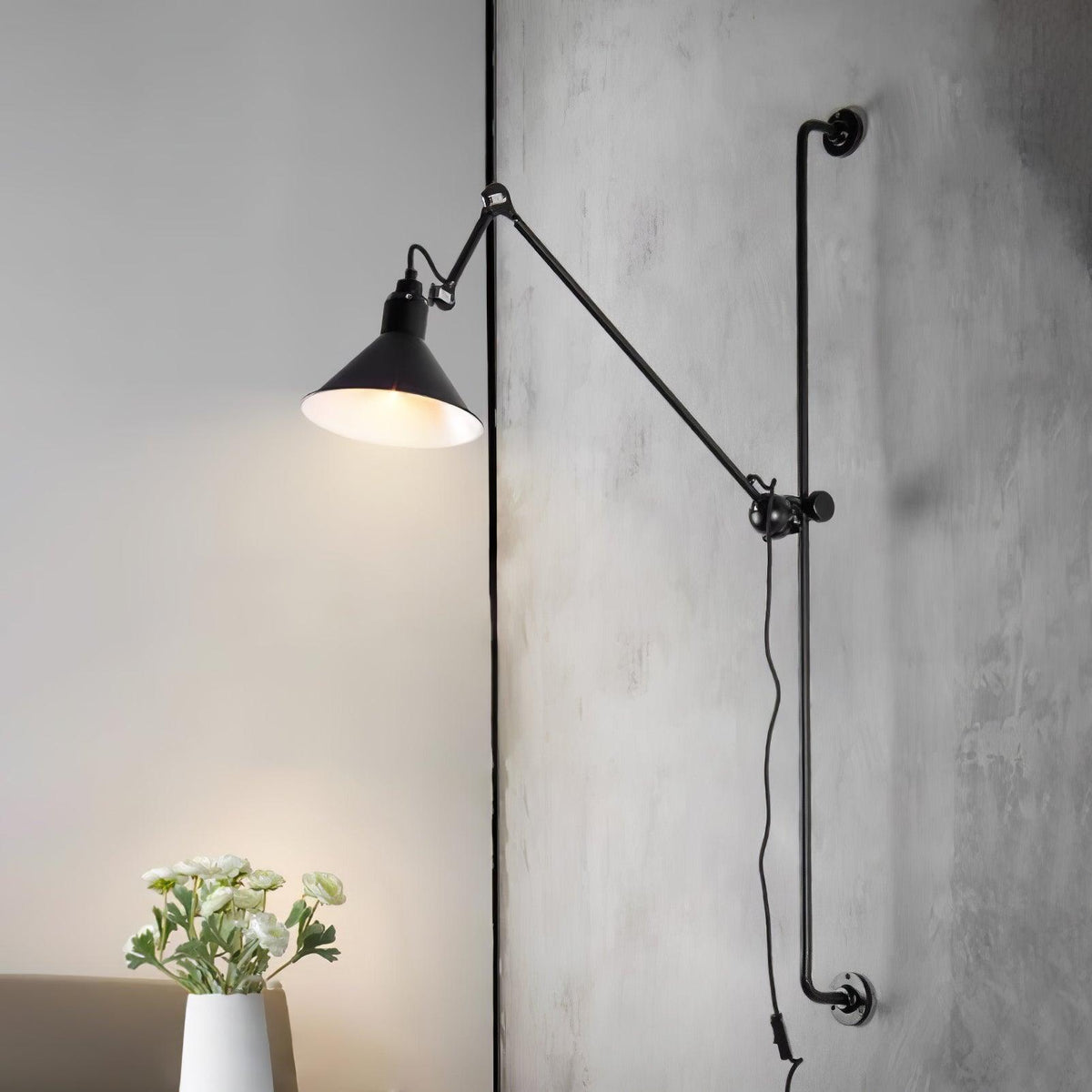 A Clérachic black wall light with a lit cone shade, mounted on a textured concrete-look wall with a white flower vase below.