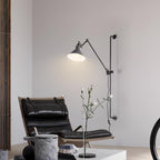 A Clérachic wall light with a white cone shade on a long black bar, beside a black leather lounge chair and a slim vase of flowers.