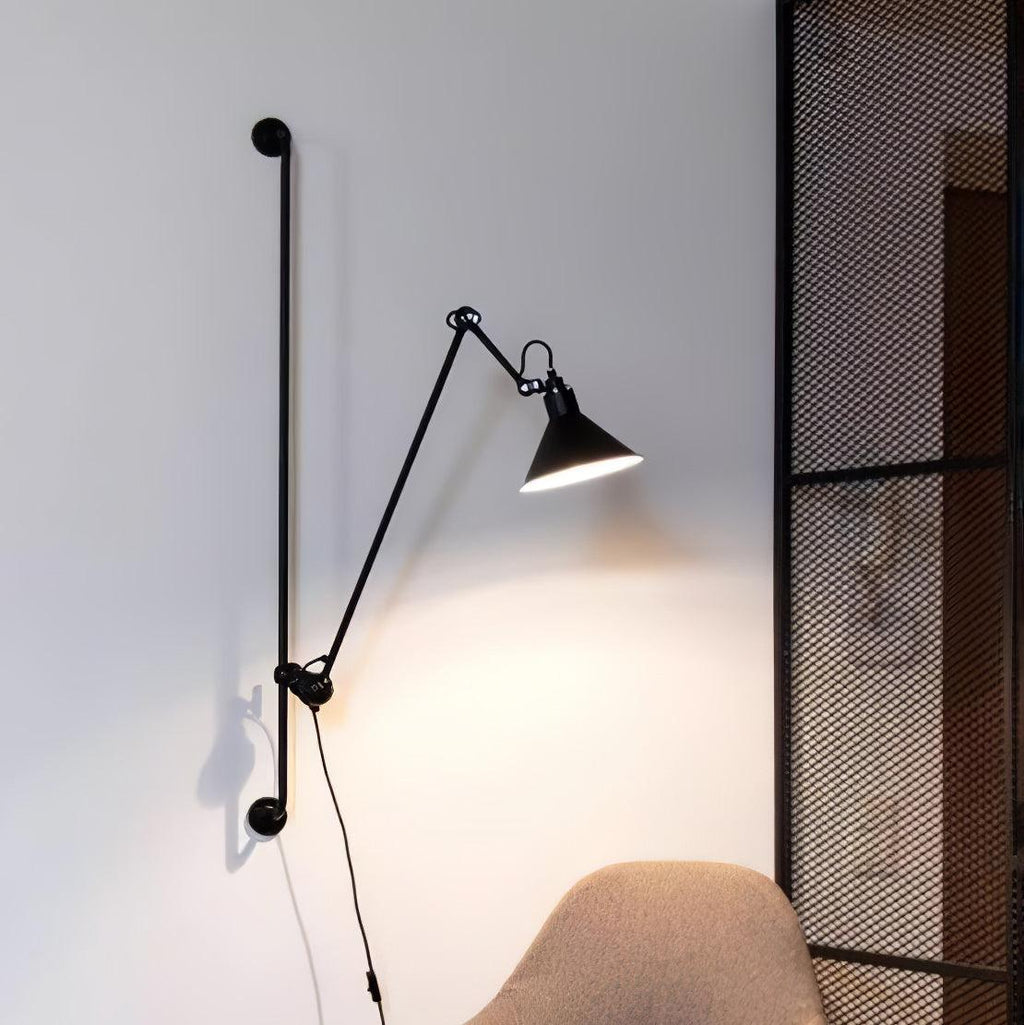 A Clérachic black wall light with a jointed arm reaching out over a light wall, with a woven chair and a metal mesh screen beside it.