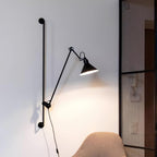 A Clérachic black wall light with a jointed arm reaching out over a light wall, with a woven chair and a metal mesh screen beside it.