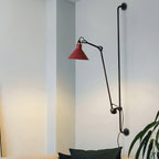 This Clérachic wall light has a red cone shade on a long black bar, lighting a pale corner beside a large leafy plant.