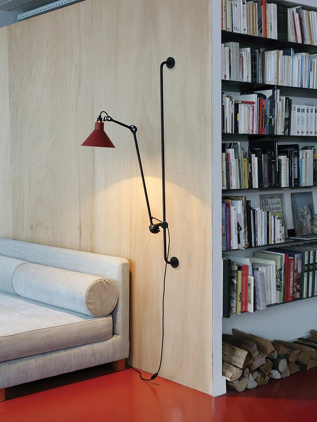Shown here, a Clérachic wall light with a red cone shade glowing against a pale wood wall, beside a tall bookshelf and a grey daybed.