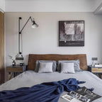 A Clérachic wall light with a pale cone shade above a bedside, on the wall beside a framed monochrome print and a wooden headboard.