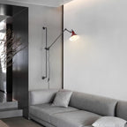 Shown here, a Clérachic wall light with a red cone shade reaching out above a grey sofa in a minimal living room with a dark panelled wall.