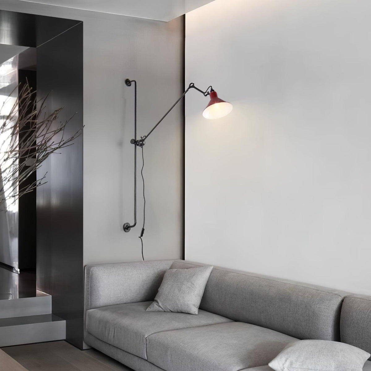 Shown here, a Clérachic wall light with a red cone shade reaching out above a grey sofa in a minimal living room with a dark panelled wall.