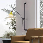 A Clérachic wall light with a yellow cone shade next to a tall window with palm leaves, above a tan leather armchair.