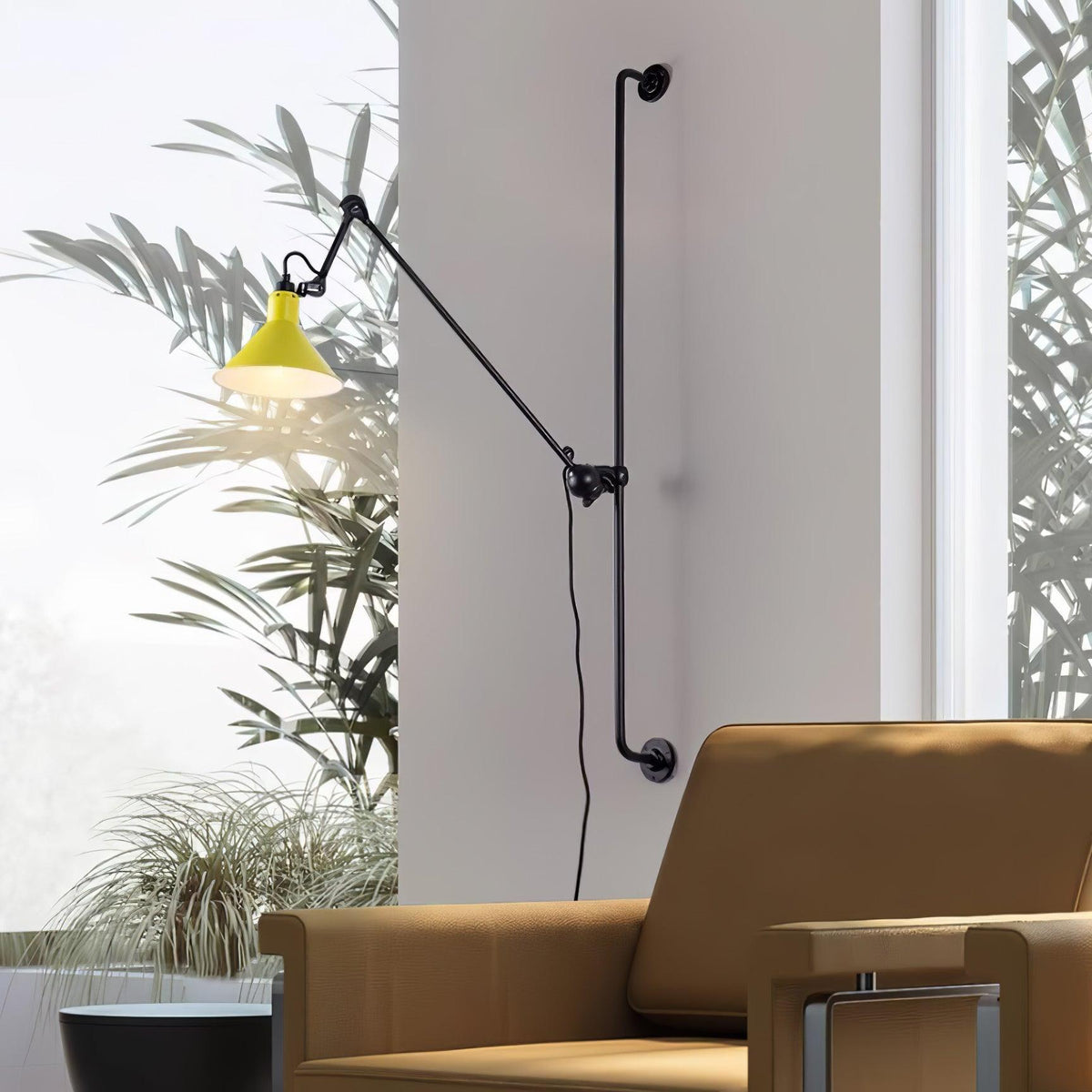 A Clérachic wall light with a yellow cone shade next to a tall window with palm leaves, above a tan leather armchair.