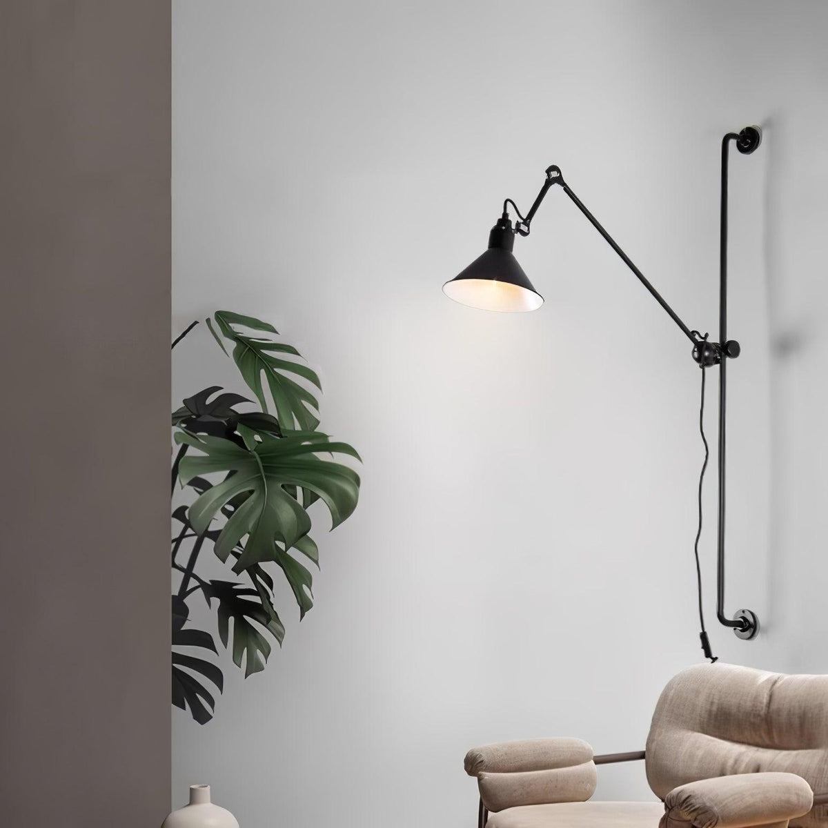 A Clérachic black wall light with a lit cone shade, beside a large monstera plant on a pale wall above a cream armchair.