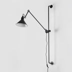 This Clérachic wall light on a plain white wall, with a long black bar, a hinged arm and a black cone shade with a white inner face.