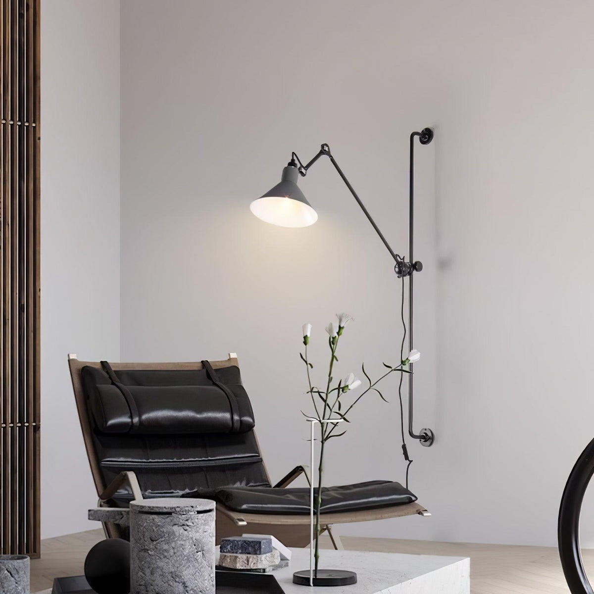 This Clérachic wall light has a white cone shade lit above a black leather lounge chair, with a stone side table and a flower stem.