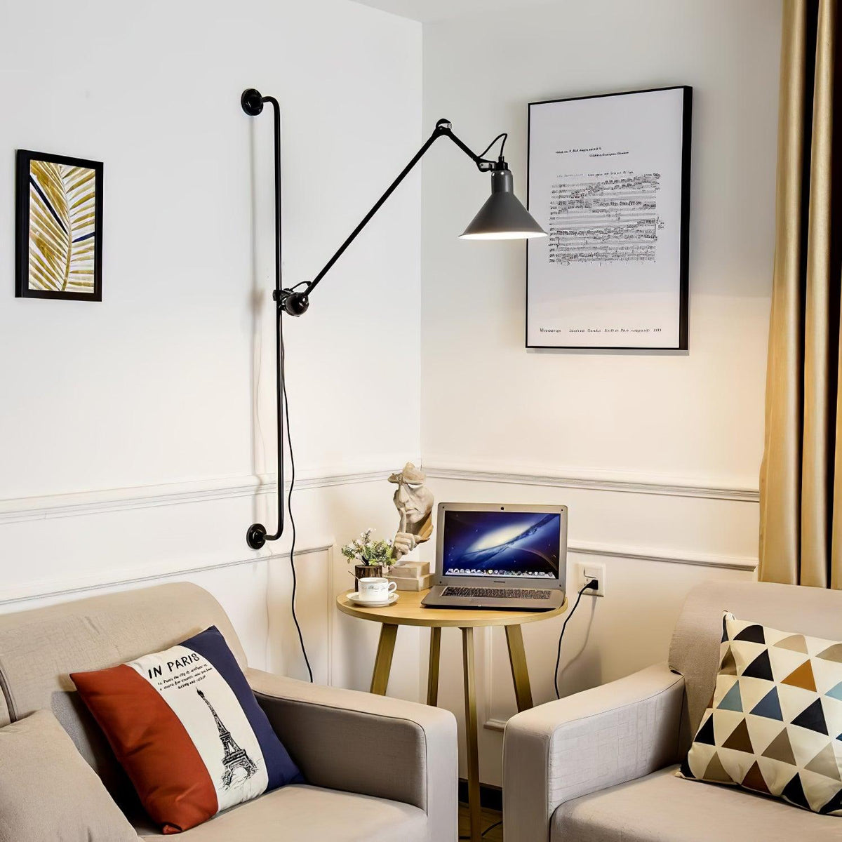 A Clérachic black wall light with a long arm across a corner of a cream room, with framed prints and a laptop on a small round table.