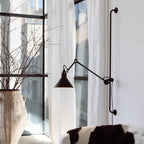 A Clérachic black wall light with a long arm and cone shade beside tall windows with white curtains and bare branches in a vase.