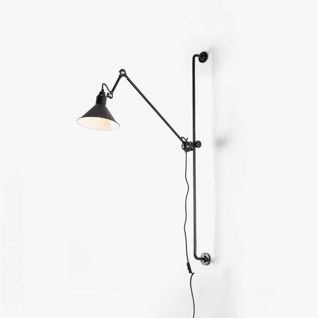 A Clérachic black wall light on a plain white wall, with the lit cone shade tilted to one side and its cable hanging below.