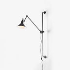 A Clérachic black wall light on a plain white wall, with the lit cone shade tilted to one side and its cable hanging below.