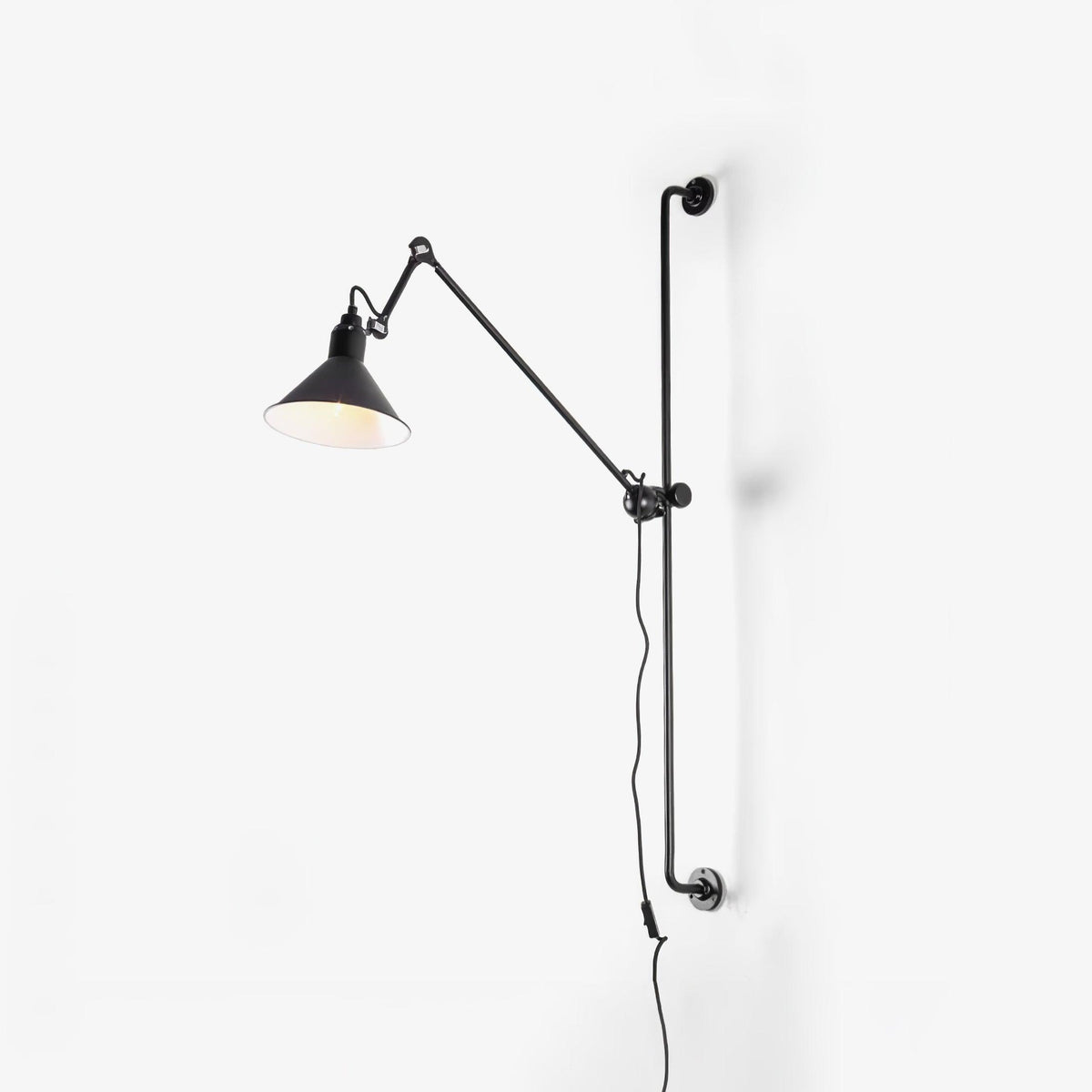 A Clérachic black wall light on a plain white wall, with the lit cone shade tilted to one side and its cable hanging below.