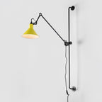 This Clérachic wall light has a yellow cone shade and a white inner face, on a long black bar against a pale grey backdrop.