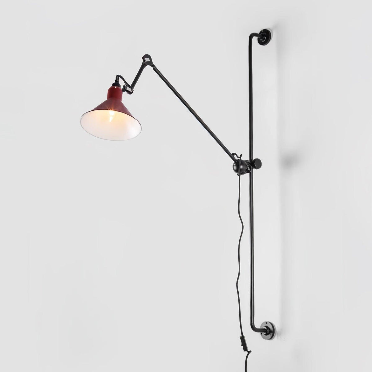 Shown here, a Clérachic wall light with a deep red cone shade on a long black bar, shown against a pale grey backdrop.