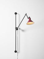 This Clérachic wall light has a deep red cone shade, its arm angled to the right, on a pale wall with the bar running down to the left.