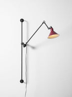 Shown here, a Clérachic wall light with a deep red cone shade swung to the right, on a pale wall with its black bar below.