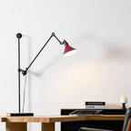 A Clérachic wall light with a deep red shade lighting the wall above a wooden desk, with a stack of books and a tablet below.