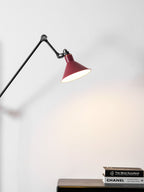 Close view of the deep red cone shade on this Clérachic wall light, lit above a stack of books on a dark sideboard.