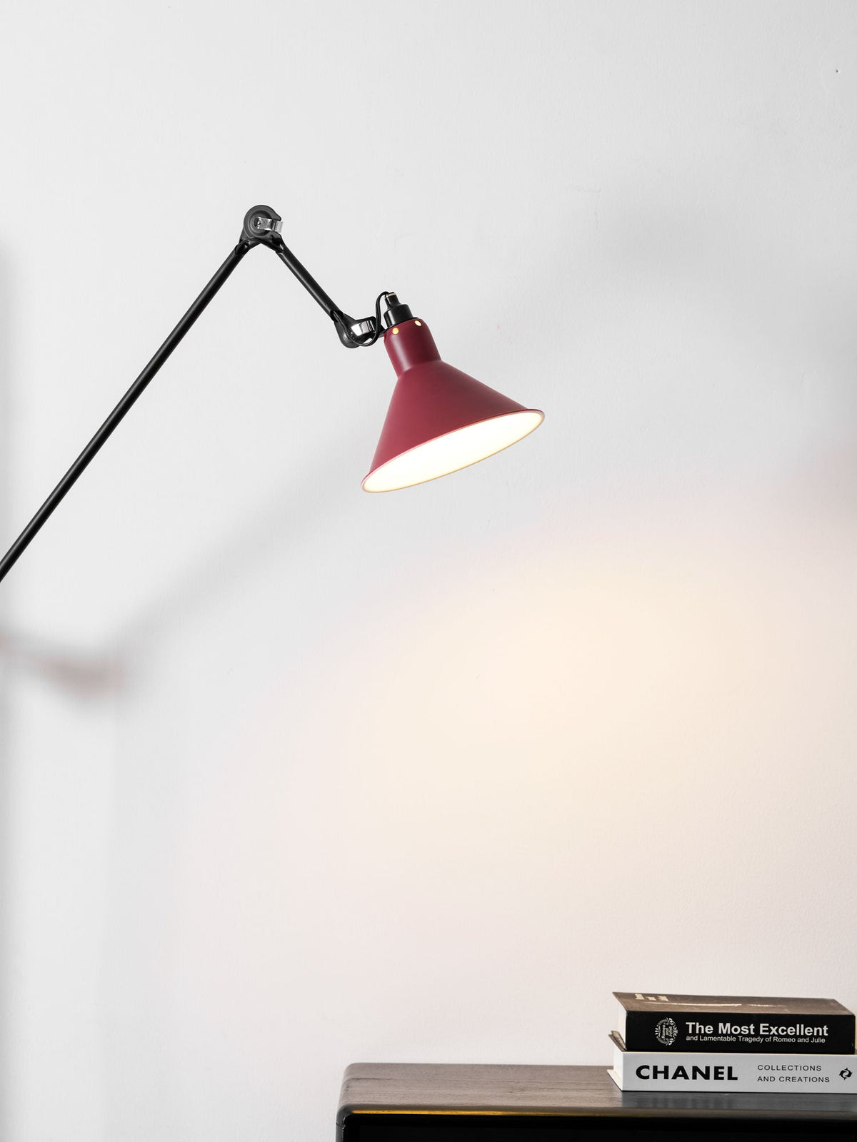 Close view of the deep red cone shade on this Clérachic wall light, lit above a stack of books on a dark sideboard.