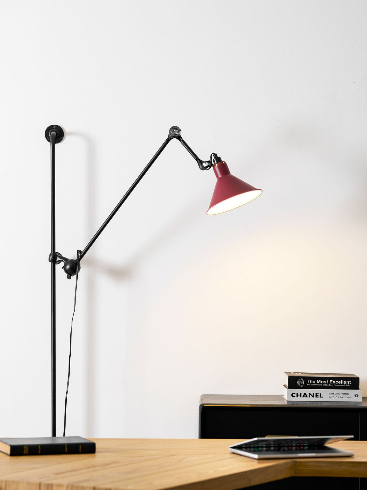 This Clérachic wall light has a red shade hung over a wooden desk, with a small stack of books and a tablet on top.