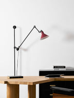 This Clérachic wall light has a red cone shade lit above a wooden desk, with a glass vase, an open book and a black cabinet.