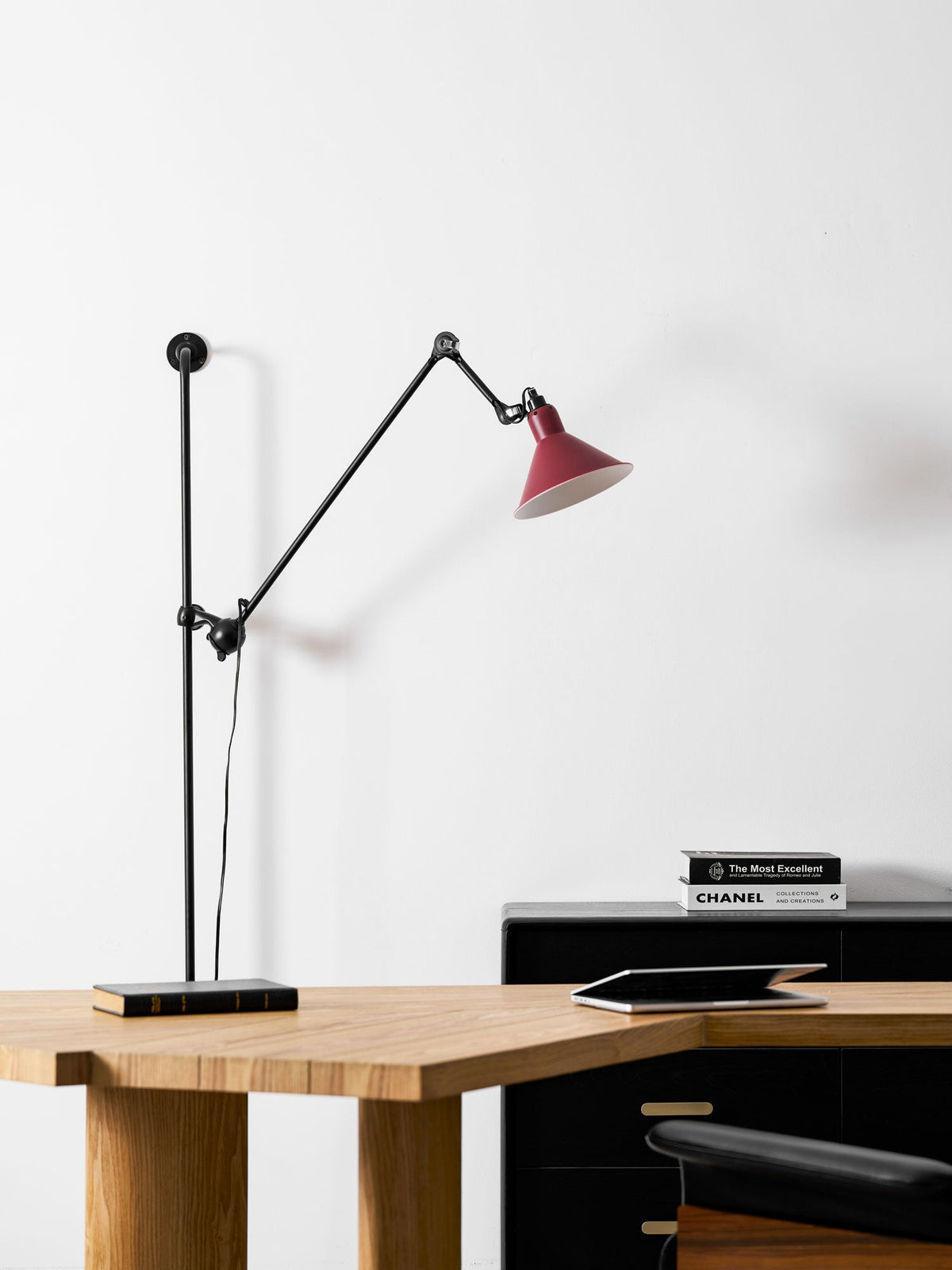 This Clérachic wall light has a red cone shade lit above a wooden desk, with a glass vase, an open book and a black cabinet.