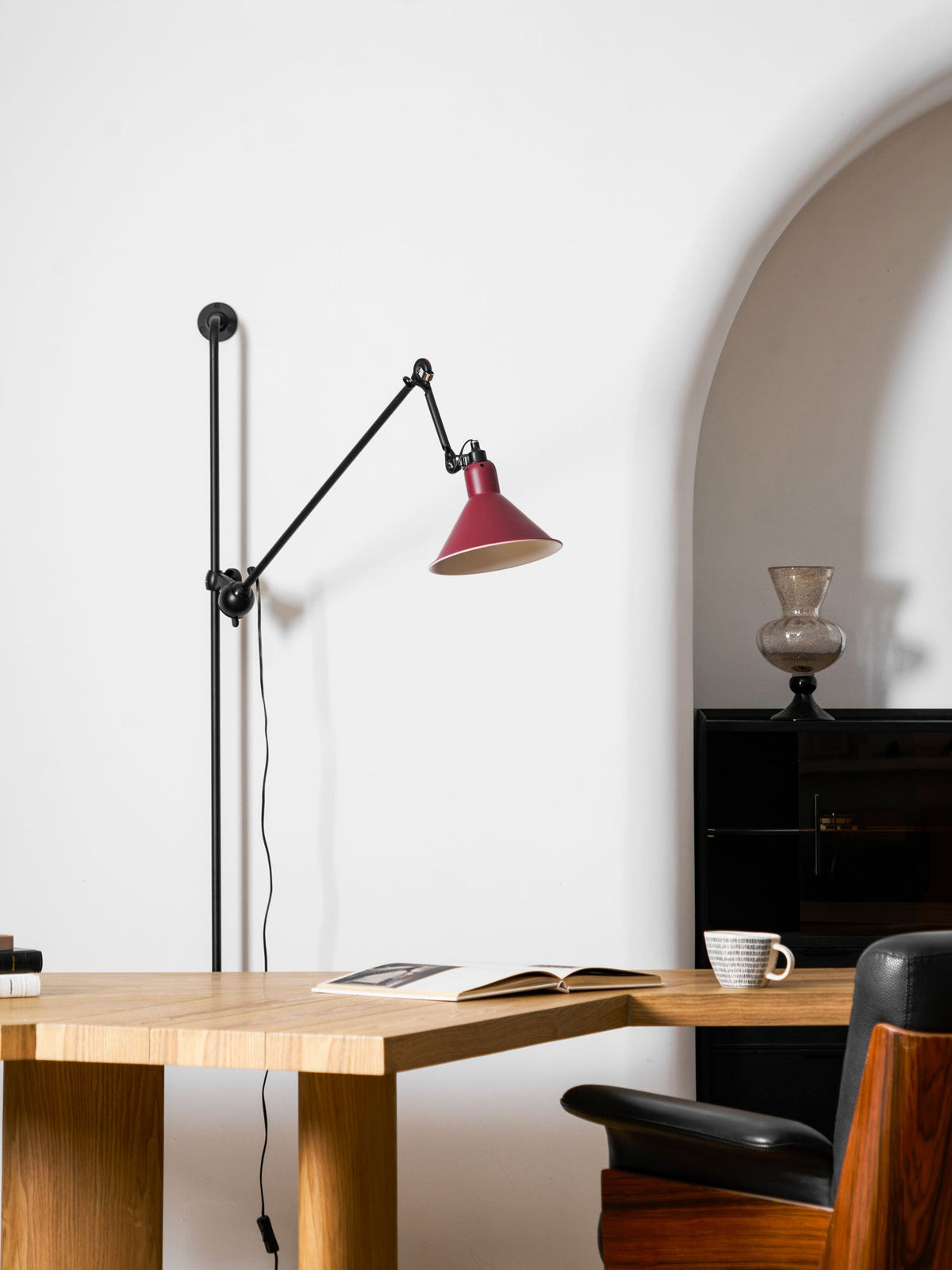 Shown here, a Clérachic wall light with a red cone shade against a white wall, with an arched niche, a desk with an open book and a tea cup.