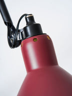 This Clérachic wall light has a red cone shade angled out from a long black bar on a pale wall, with the cable hanging down.