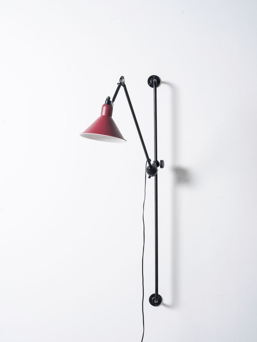 Looking up at this Clérachic wall light, a red cone shade with a warm bulb inside and a hinged black arm above it.