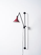 Looking up at this Clérachic wall light, a red cone shade with a warm bulb inside and a hinged black arm above it.