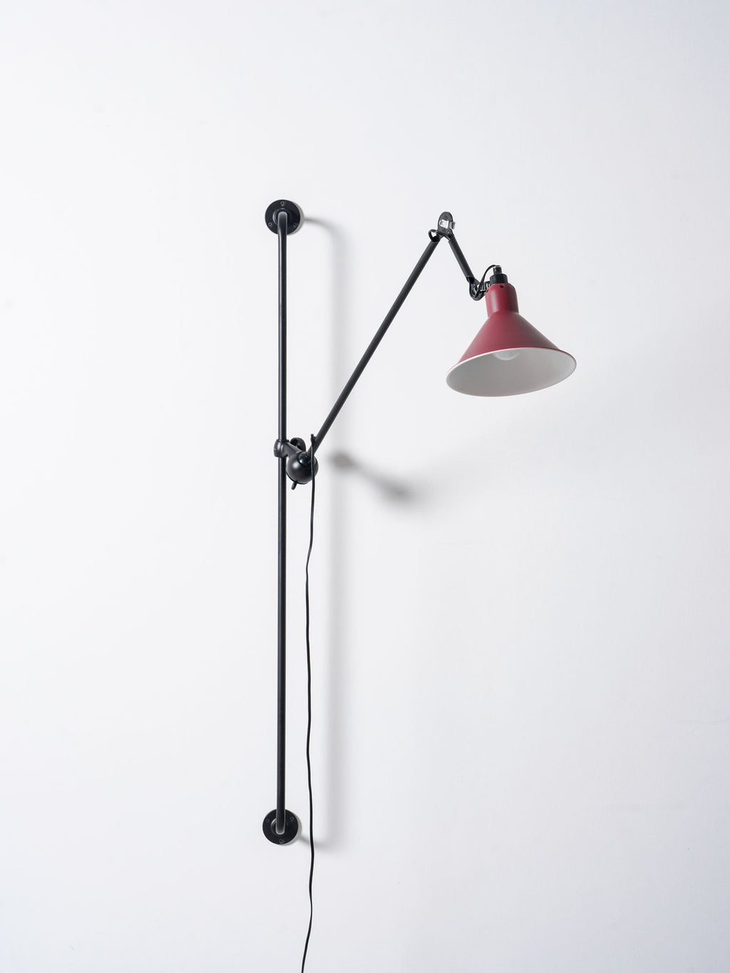 A Clérachic wall light with the red cone shade tilted down, the black arm folded back toward the bar on a plain pale wall.