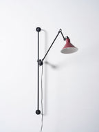 A Clérachic wall light with the red cone shade tilted down, the black arm folded back toward the bar on a plain pale wall.