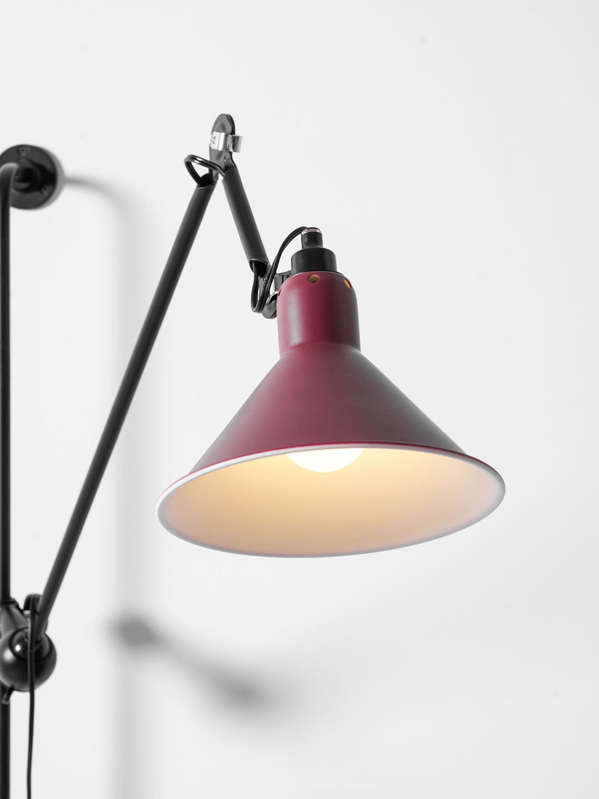 A Clérachic wall light close up, with a red cone shade, a lit bulb inside it and a black hinged arm against a pale wall.