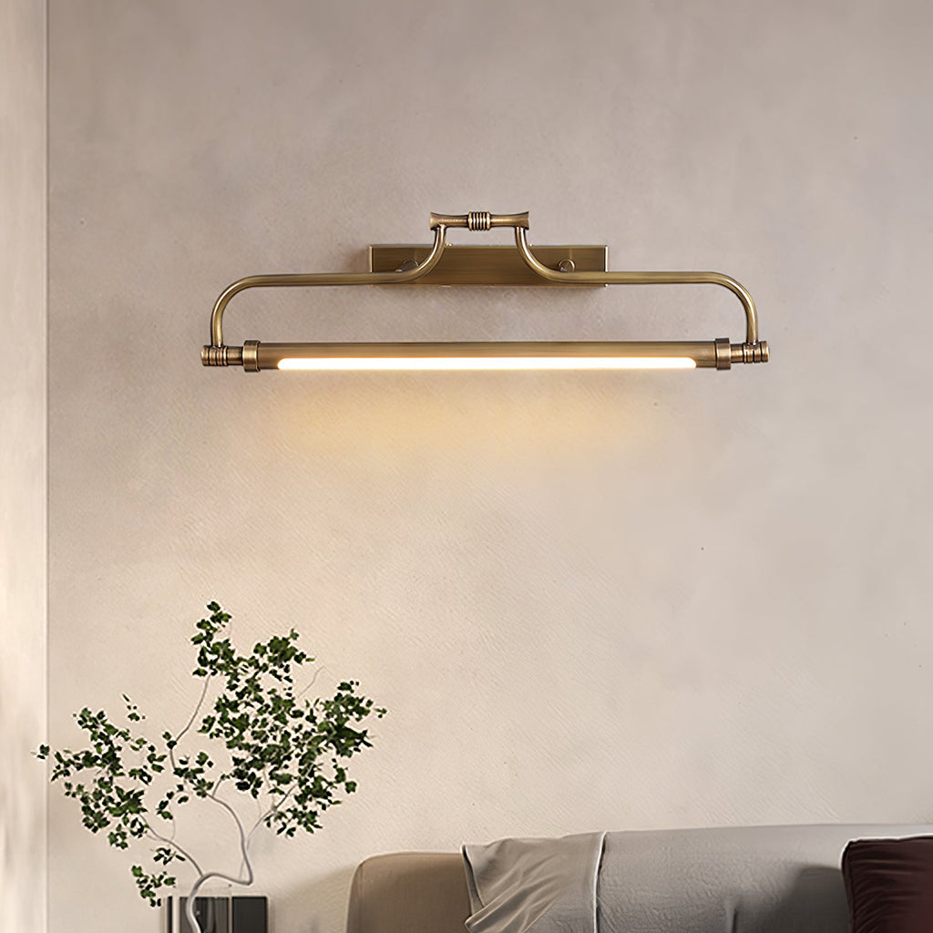 A lit Clérachic wall light on a soft beige wall, with a leafy branch in the lower corner and a grey bed edge below.