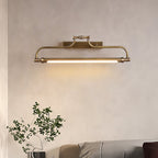 A lit Clérachic wall light on a soft beige wall, with a leafy branch in the lower corner and a grey bed edge below.