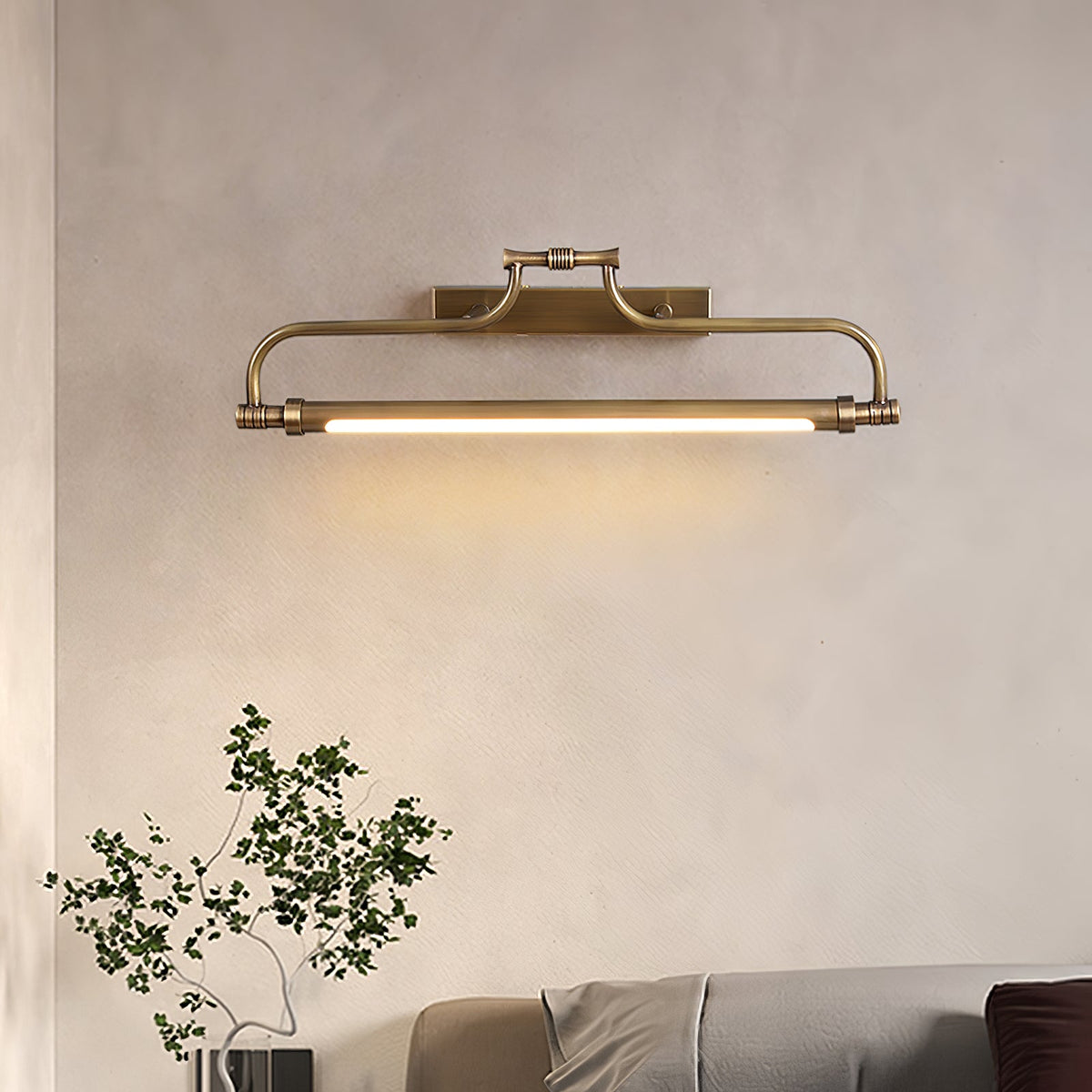 A lit Clérachic wall light on a soft beige wall, with a leafy branch in the lower corner and a grey bed edge below.
